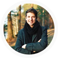 Smiling young man in dark coat and scarf standing with arms crossed outdoors in a forest setting.