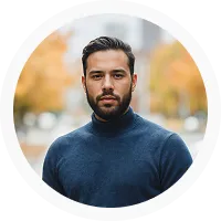 Man with dark hair and beard wearing a navy blue sweater, with a blurred background of trees and buildings.